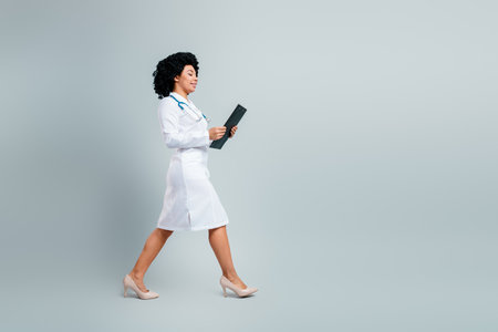 Confident young female doctor walking with clipboard in hand wearing white medical uniformの写真素材