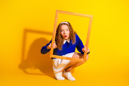 Young woman holding wooden frame against bright yellow background expressing surprise with vibrant fashion and styleの写真素材