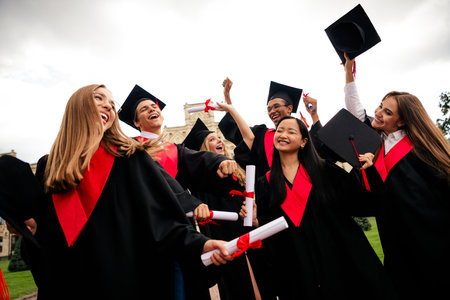 Joyful young graduates celebrating together outdoors, wearing black mantles and mortarboards,...