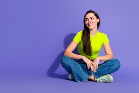 Smiling young woman sitting cross legged on bright purple background wearing a neon green shirt and jeansの写真素材