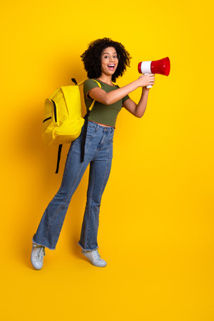 Happy young woman holding a megaphone and wearing a backpack on a vibrant yellow background, expressing excitementの写真素材