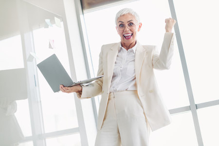 Confident senior businesswoman celebrating success at work in an office holding laptop in elegant...