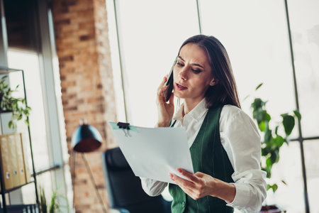 Busy businesswoman reviewing documents in a modern office while talking over the phone for work communicationの写真素材