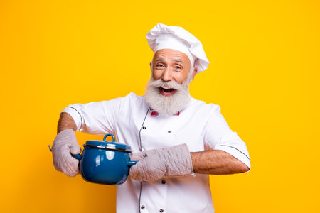 Happy chef holding a blue pot against a bright yellow background, wearing traditional uniform and displaying enthusiasm for cookingの写真素材