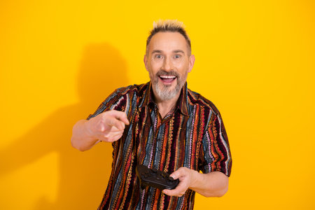 Happy mature man with a stylish look enjoys gaming against a bright yellow background demonstrating...