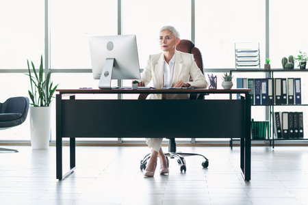 Mature professional businesswoman in modern office workspace seated at computer desk focused and confidentの写真素材