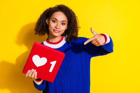 Young woman pointing at a gift box with a heart sign symbol, yellow background and casual sweater...
