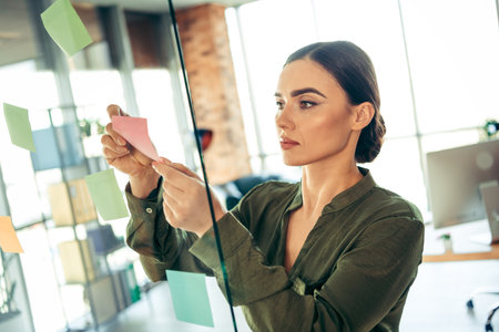 Focused businesswoman organizing her tasks using sticky notes in a modern office environment during...