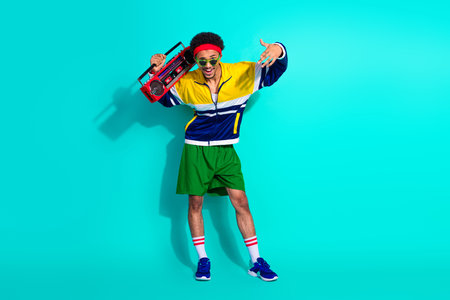 Energetic young man in retro sportswear posing with a boombox against a vibrant turquoise...