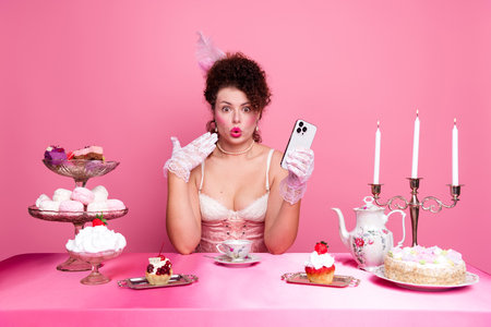 Young woman in costume enjoying pastries at a pink-themed tea party, capturing the fusion of aristocratic fashion and modern technologyの写真素材