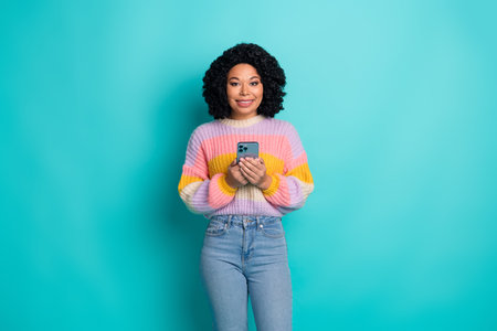 Smiling young woman holding smartphone on turquoise background, wearing colorful sweater and jeans,...