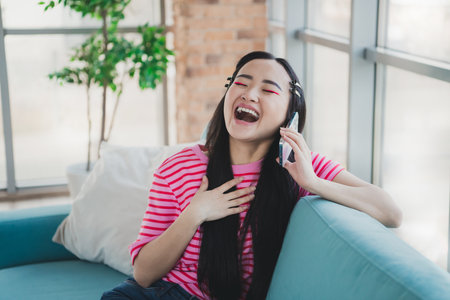 Young woman laughing while talking on smartphone, sitting on couch in cozy living room, enjoying relaxing moment during daytimeの写真素材