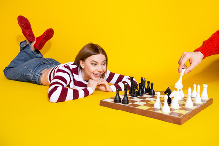 Smiling young woman enjoying a chess game on a yellow background, wearing a striped pullover and trendy jeansの写真素材