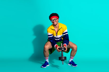 Young guy with vibrant sportswear lifting a dumbbell on an isolated teal background showcasing retro style and energyの写真素材