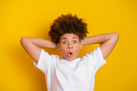 A surprised boy in a white t-shirt expresses amazement with his hands behind his head against a vibrant yellow background.の写真素材
