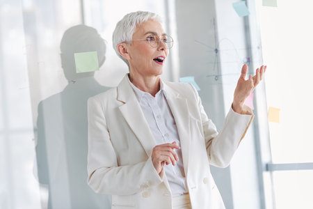 Confident senior businesswoman sharing ideas in a modern office with clear glass walls and colorful notes on boardの写真素材