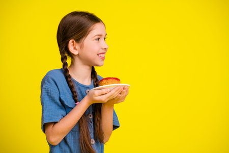 Smiling young girl in casual blue outfit holding a plate with food on a vibrant yellow backgroundの写真素材