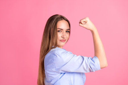 Confident young woman showing strength with flexed arm on pink background, representing empowerment, determination, and self-confidenceの写真素材