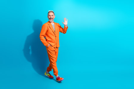 Stylish middle aged man in orange suit waving on blue background showcasing charisma and happinessの写真素材
