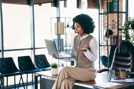 Confident businesswoman engaging in an office video meeting on her laptop while sitting on a desk in a modern workspace.の写真素材