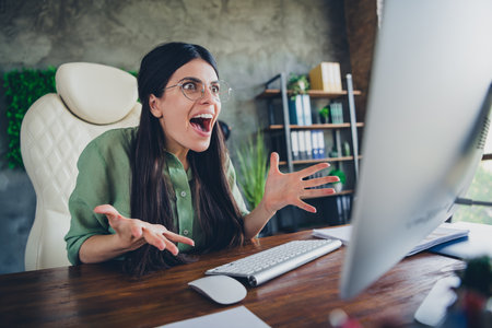 Excited young woman in formal attire expressing joy at office desk in a bright workspaceの写真素材