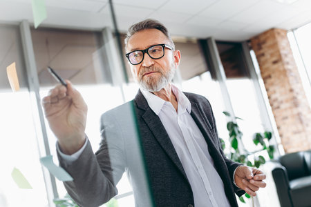 Mature business professional writing on transparent board during a brainstorming session in a well-lit modern office environmentの写真素材