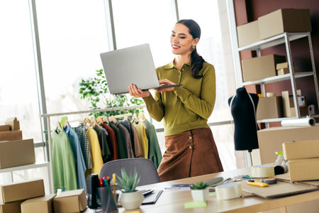 Young professional female designer using a laptop in a modern atelier surrounded by fashion designs and textilesの写真素材