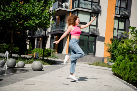 Young woman with curly hair joyfully leaps in the air, wearing a trendy pink top and jeans, enjoying a sunny day in an urban settingの写真素材