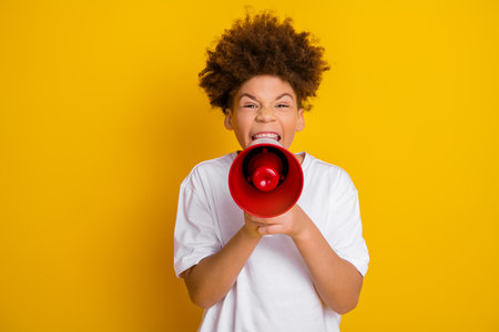 Cheerful boy with curly hair using a bright red megaphone against a vibrant yellow backgroundの写真素材