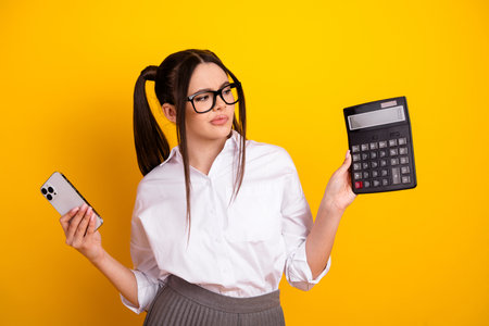 Young female student in stylish attire holding a smartphone and calculator against a bright yellow backgroundの写真素材