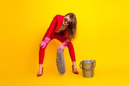Stylish young woman in red suit and pink gloves having fun cleaning with mop on bright yellow backgroundの写真素材