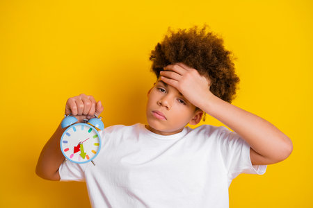 Boy holding an alarm clock looking thoughtful in front of a bright yellow background expressing...