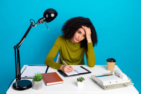 Thoughtful young woman writing at desk in bright workspace with blue background and office items...