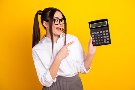 Young female student in a white shirt holding a calculator and pointing, cheerful expression on a yellow backgroundの写真素材