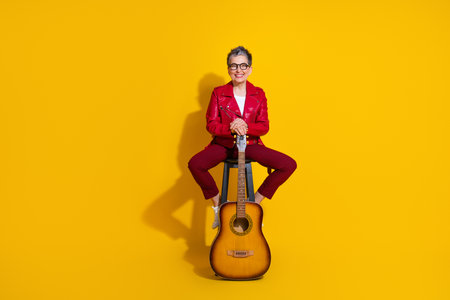 Stylish elderly woman in red jacket holding a guitar on a vibrant yellow background with charming...