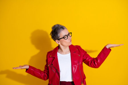 Stylish mature woman in vibrant red jacket posing with playful hand gestures against bright yellow...