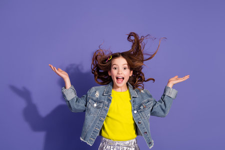 Joyful preteen girl in trendy denim jacket smiling with hands raised against a vibrant purple backgroundの写真素材