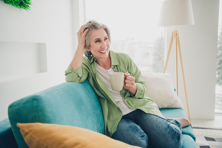 Charming mature woman enjoying leisure time at home with coffee on a cozy sofa in a bright living roomの写真素材