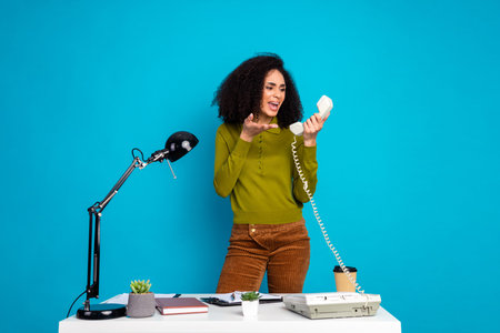 Young woman in stylish outfit holding retro telephone handset while gesturing enthusiastically at desk in vibrant blue settingの写真素材