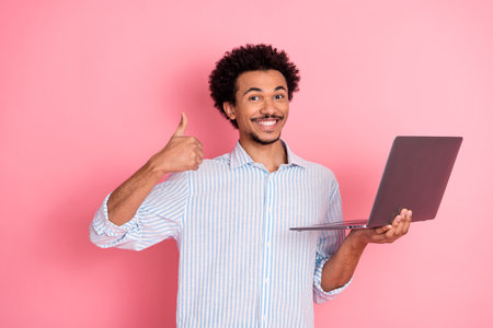 Confident young man holding laptop and giving thumbs up on pink background promoting business,...