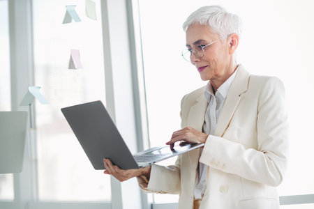 Successful senior businesswoman working with a laptop in a bright modern office space, showcasing professionalism and determinationの写真素材