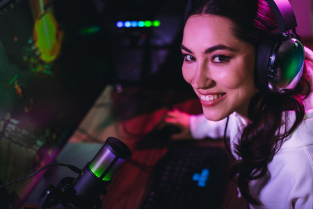 Young female gamer livestreaming video gameplay with a computer and microphone in a neon-lit gaming...