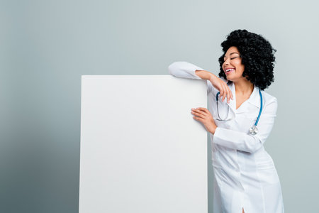 Confident female doctor with a stethoscope leaning on a blank board against a grey background, smiling brightlyの写真素材