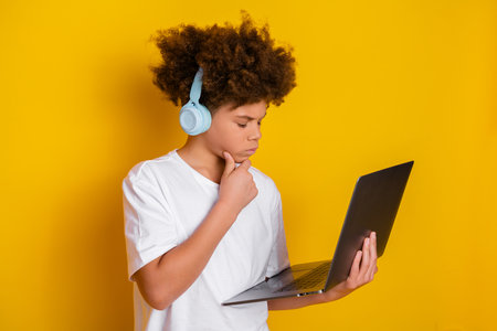 Young boy wearing headphones and holding a laptop on a vibrant yellow background, pondering and...