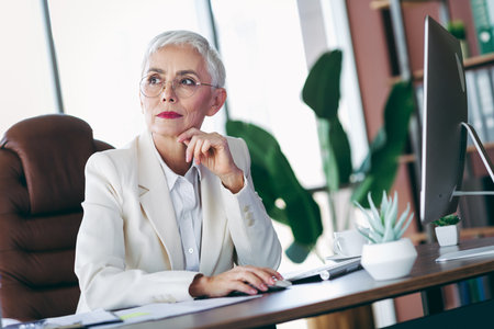 Elegant mature businesswoman with short white hair sitting at a desk in a modern office workspace, looking reflectiveの写真素材