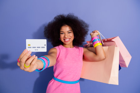 Stylish young woman with afro hairstyle holding shopping bags and a credit card, dressed in a bright...