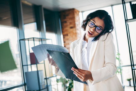 Confident businesswoman multitasking in a modern office holding documents and using a smartphone for communicationの写真素材