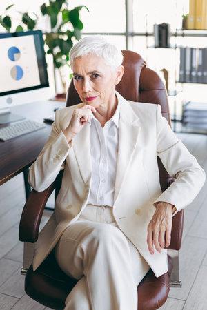 Sophisticated mature businesswoman in white suit sitting confidently in office chairs, demonstrating professionalism and leadershipの写真素材