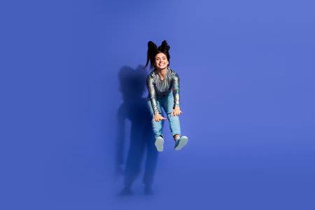 Stylish young woman in glossy top jumping joyfully on a bright violet background, showcasing...
