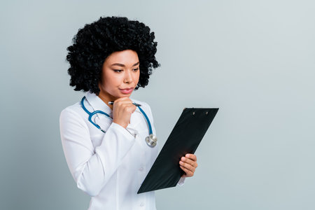 Female physician in white coat examining patient file against light background, representing healthcare professionalismの写真素材